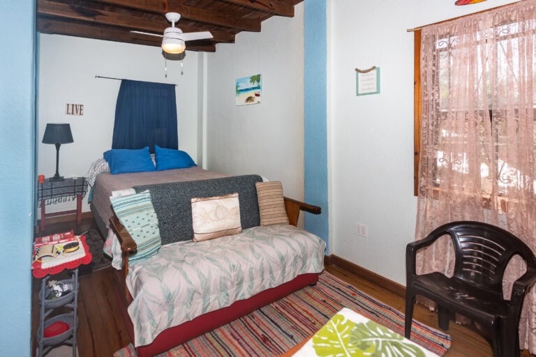 A cozy bedroom in a Roatan vacation rental featuring a bed with beach-themed pillows, a ceiling fan, colorful wall art depicting the ocean, and pink lace curtains allowing natural light into the space Honduran style.