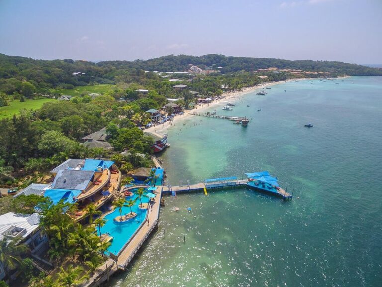 Aerial view of a popular beachfront resort in Roatan, Honduras, featuring a large swimming pool surrounded by palm trees, colorful buildings, and a pier extending into the turquoise Caribbean Sea.