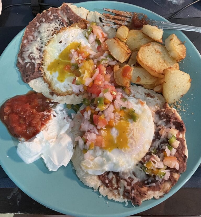 A plate of traditional Honduran baleadas served on a turquoise dish in Roatan, Honduras, featuring refried beans, eggs, fresh tomato and onion salsa, sliced potatoes, and optional hot sauce.