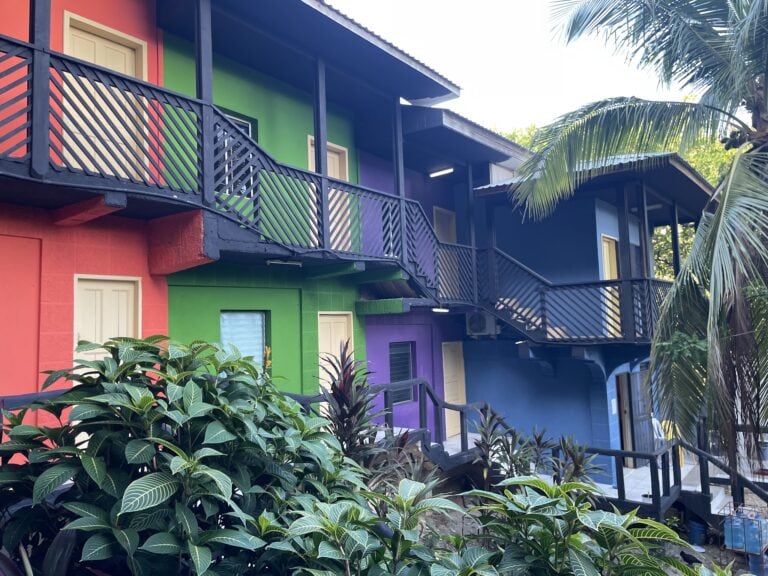 A colorful multi-story building with green, purple, and black exterior walls, featuring balconies with black railings and surrounded by lush tropical foliage on Roatan, Honduras.
