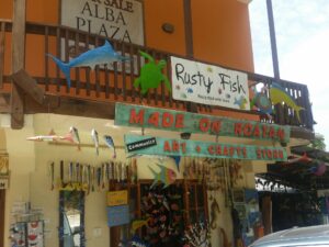 Colorful fishing-themed crafts and signs, including a "Rusty Fish" recycled art display, are shown on a wooden storefront in Roatan, Honduras, promoting local art and sustainability.