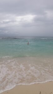 A person swimming in turquoise waters near a sandy beach under a cloudy sky on Roatan in Honduras.