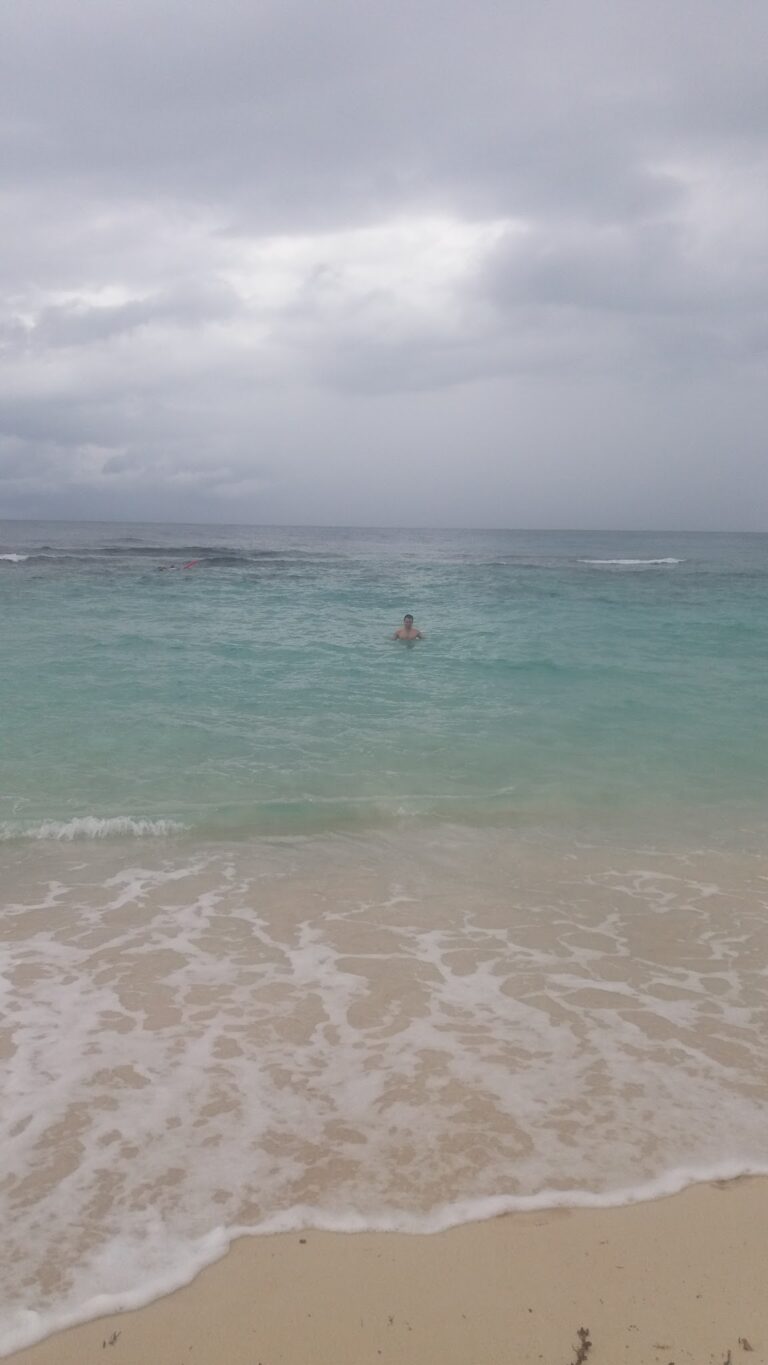 A person swimming in turquoise waters near a sandy beach under a cloudy sky on Roatan in Honduras.