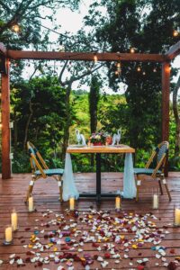 A romantic outdoor dining setup on a wooden deck in Roatan, Honduras, features a table decorated with a flower arrangement, swan-shaped centerpieces, surrounded by candles and rose petals, with lush green trees and string lights overhead creating a cozy, intimate atmosphere.