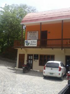 A two-story building on Roatan, Honduras, features a yellow exterior, a balcony with a wooden railing, and a sign indicating it is for sale at Alba Plaza; the area has a cobblestone street with parked cars and motorcycles, surrounded by lush green trees.