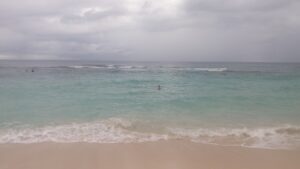 A person is swimming in the clear, turquoise waters of Roatan, Honduras, with a cloudy sky overhead and gentle waves lapping onto the sandy beach.