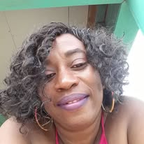 A woman with curly gray hair and hoop earrings poses for a selfie indoors against a light green wall, with the setting associated with a business listing on RoatanYP.com in Roatan, Honduras.