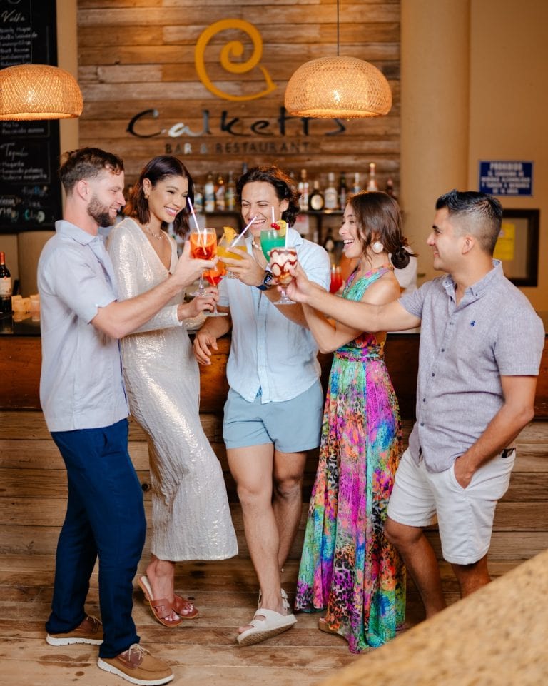 A group of six friends celebrating with colorful cocktails in a lively bar on Roatan, Honduras, standing in front of a wooden wall with the "Calkets Bar & Restaurant" sign and a collection of liquor bottles.