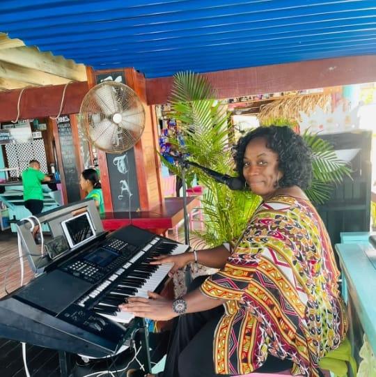 A woman playing a keyboard and singing into a microphone in an outdoor-rooftop setting with palm plants and colorful decor in Roatan, Honduras.