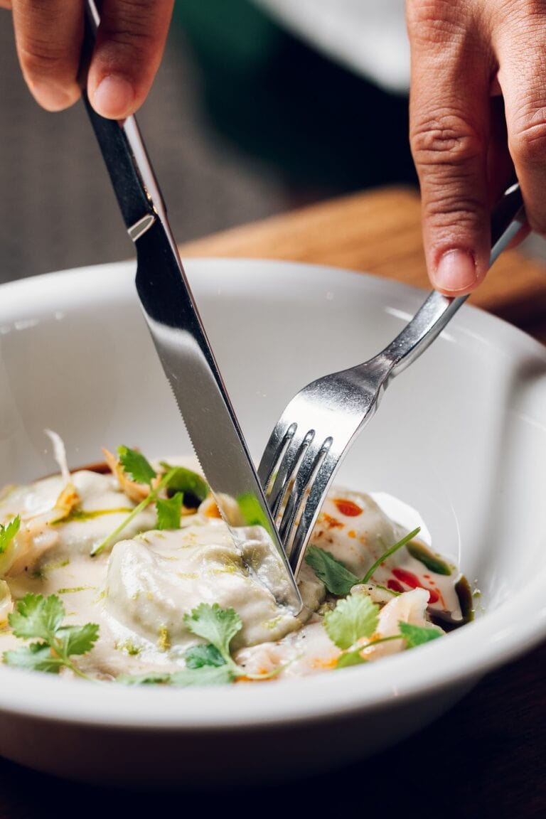 A person uses a fork and knife to cut into a creamy, garnished dish in a white bowl on a dark surface, with the dish featuring herbs, possibly cilantro, and colorful sauces, in Roatan, Honduras.