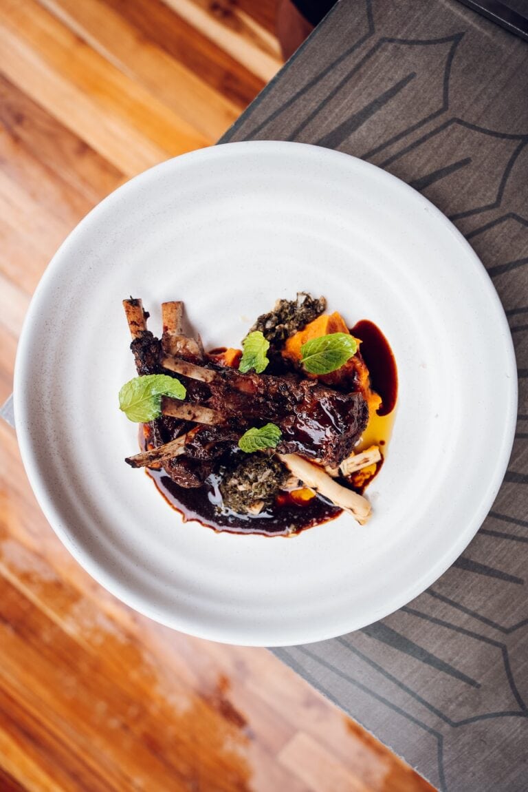 A plated dish featuring grilled lamb chops with a dark glaze, garnished with fresh green herbs, served alongside sweet potato mash and a spinach mixture, on a white ceramic plate placed on a textured gray surface in Roatan, Honduras.