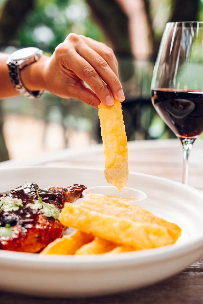 A hand holding a piece of fried cheese above a plate of food with red wine in a glass, set on a wooden table in a tropical outdoor restaurant in Roatan, Honduras.