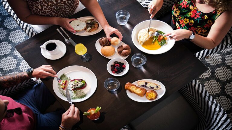 A group of people enjoying a meal at a dark wooden table in a restaurant on Roatan, Honduras, with various plates of food, desserts, beverages, and a plate of assorted bread surrounded by patterned chairs and hexagonal tile flooring.