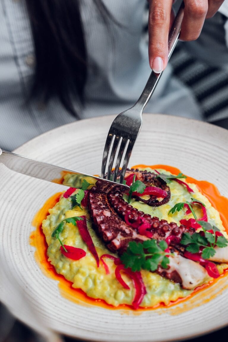 A plate of creamy mashed potatoes topped with grilled octopus, red onion, chopped herbs, and vibrant red and green sauce, served on a white ceramic dish in Roatan, Honduras.