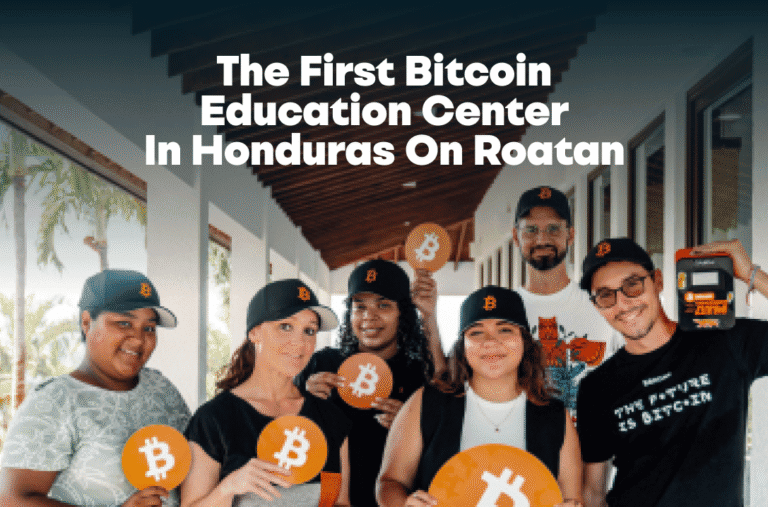 A group of people wearing black caps with Bitcoin logos and holding Bitcoin symbols pose inside a building with large windows and wooden ceiling beams, promoting the first Bitcoin education center on Roatan, Honduras.