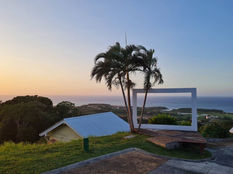 A scenic view from Roatan, Honduras, featuring palm trees, a white geometric frame on a grassy hill, and a coastal landscape with houses and the ocean in the background during sunset.
