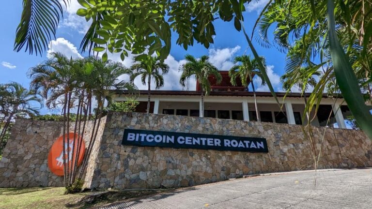 A tropical building with a stone facade and a sign reading "Bitcoin Center Roatan" is surrounded by palm trees and lush greenery, set against a bright blue sky in Roatan, Honduras.
