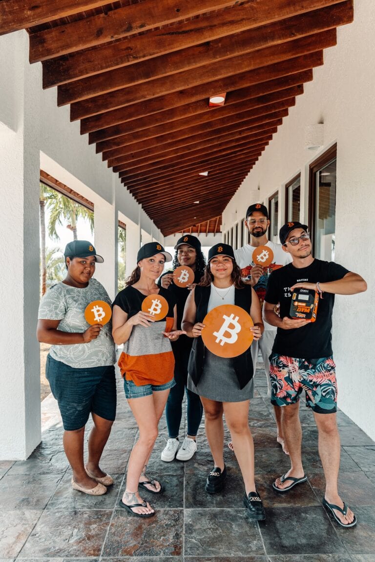 A group of six diverse people holding Bitcoin signs stand together under a wooden pergola on a patio in Roatan, Honduras, with palm trees visible outside.