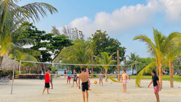 People playing volleyball on a sandy beach surrounded by palm trees and thatched-roof huts in Roatan, Honduras.