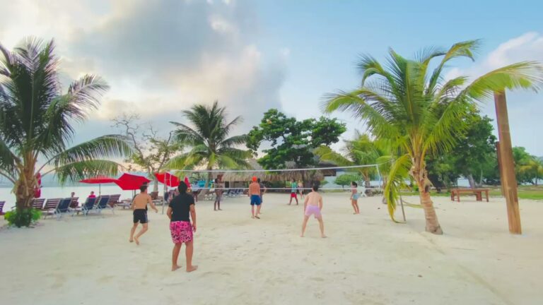 People are playing beach volleyball on a sandy shore with palm trees, lounge chairs, and umbrellas along the beachfront in Roatan, Honduras.