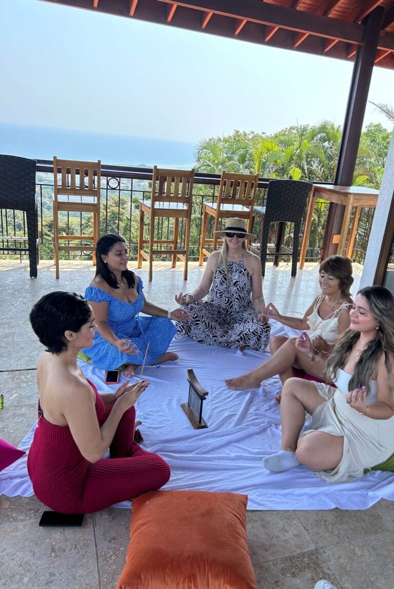 A group of women practicing meditation on a white cloth at an outdoor terrace in Roatan, Honduras, with lush greenery, palm trees, and a clear blue sky in the background.