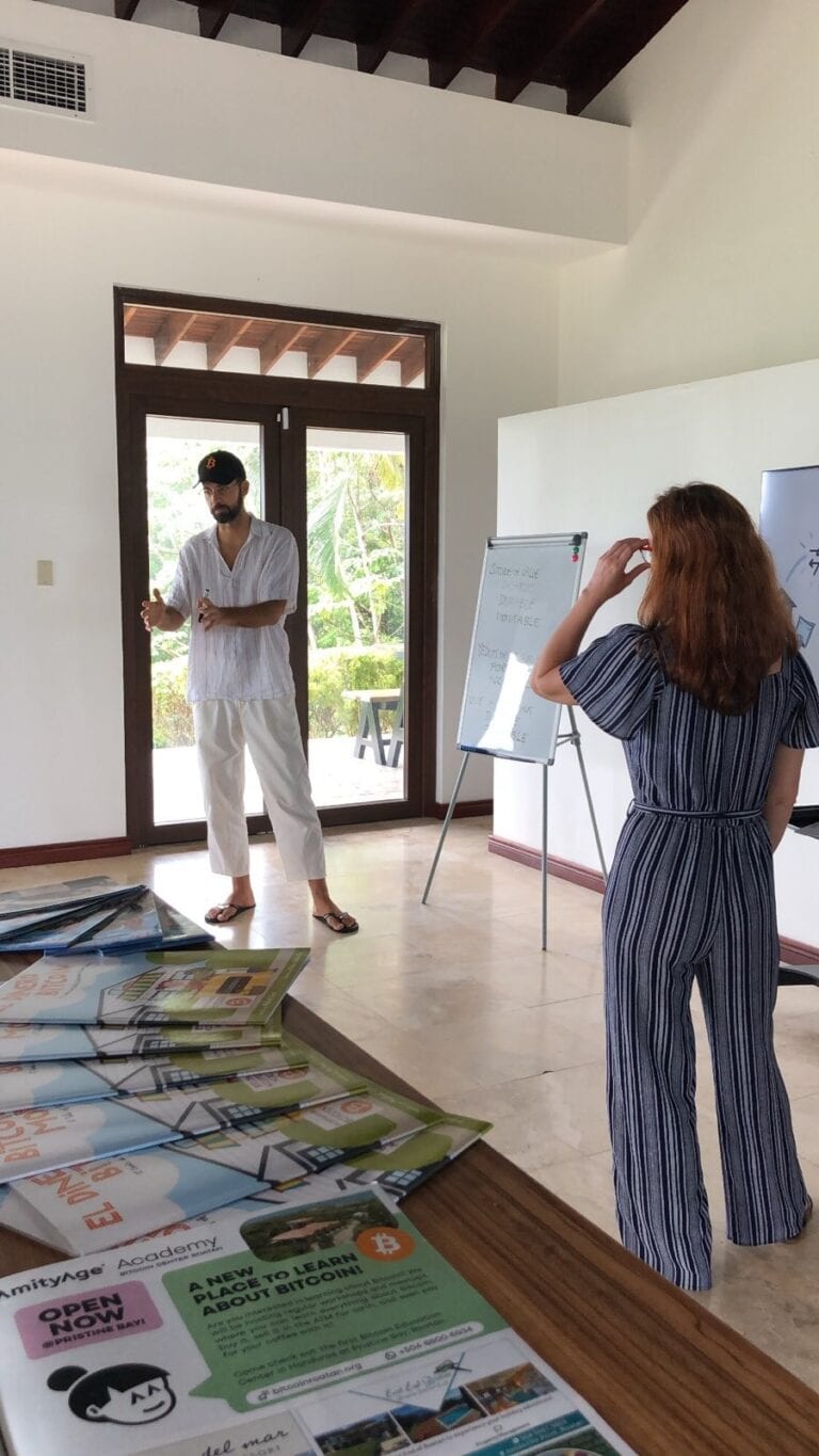 A man and a woman are having a discussion inside a bright room with large glass doors showing a lush green outdoor setting in Roatan, Honduras, while a flip chart and brochures about Bitcoin are visible nearby.