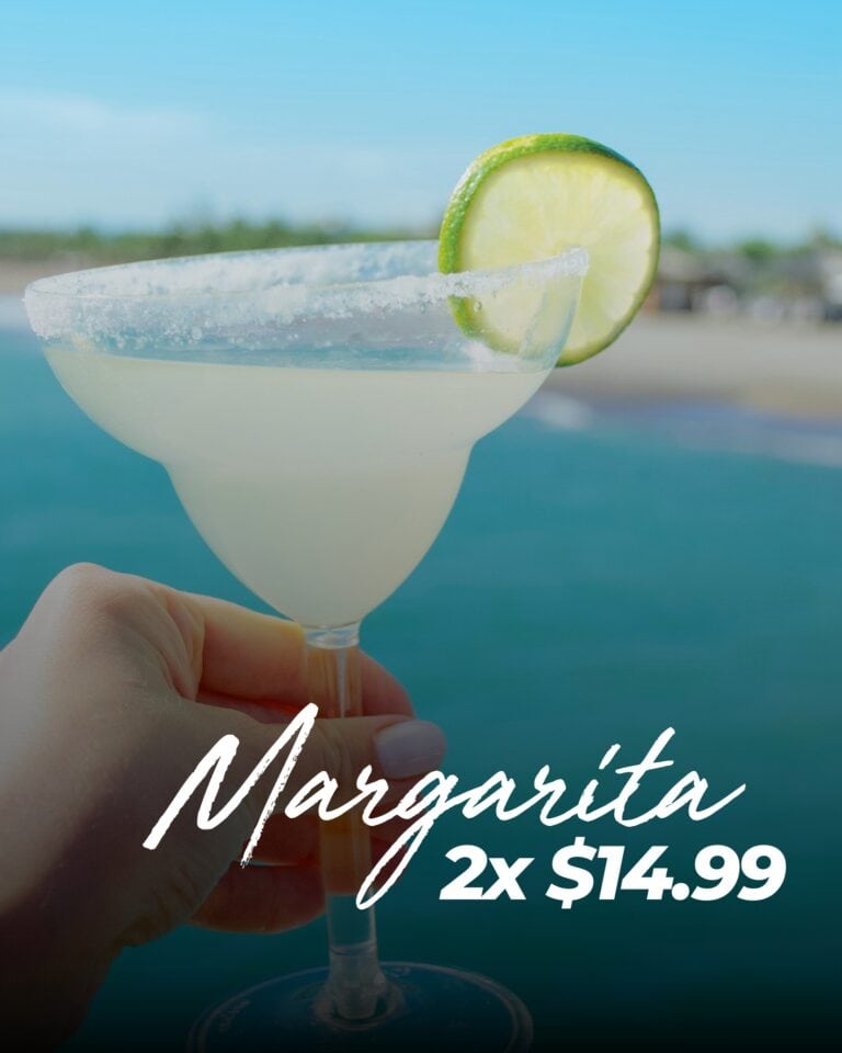 A hand holds a margarita glass with a salted rim and a lime wedge garnishing the edge against a blurred outdoor background with trees and sky in Roatan, Honduras.