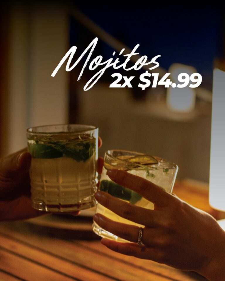 A hand with several rings holding a small glass of a layered drink with lime and mint garnishes, illuminated by warm dim lighting in a cozy setting in Roatan, Honduras.