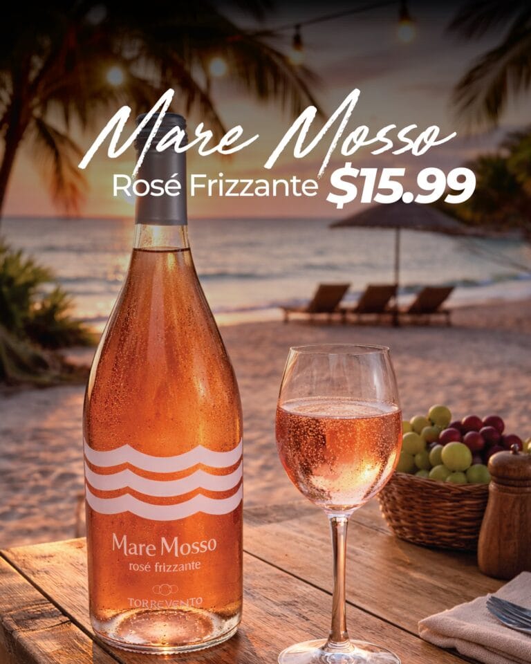 A bottle of Mare Mosso rosé frizzante sparkling wine and a glass filled with the beverage are set on a wooden table at sunset near the beach in Roatan, Honduras, with palm trees, string lights, and a thatched umbrella visible in the background.