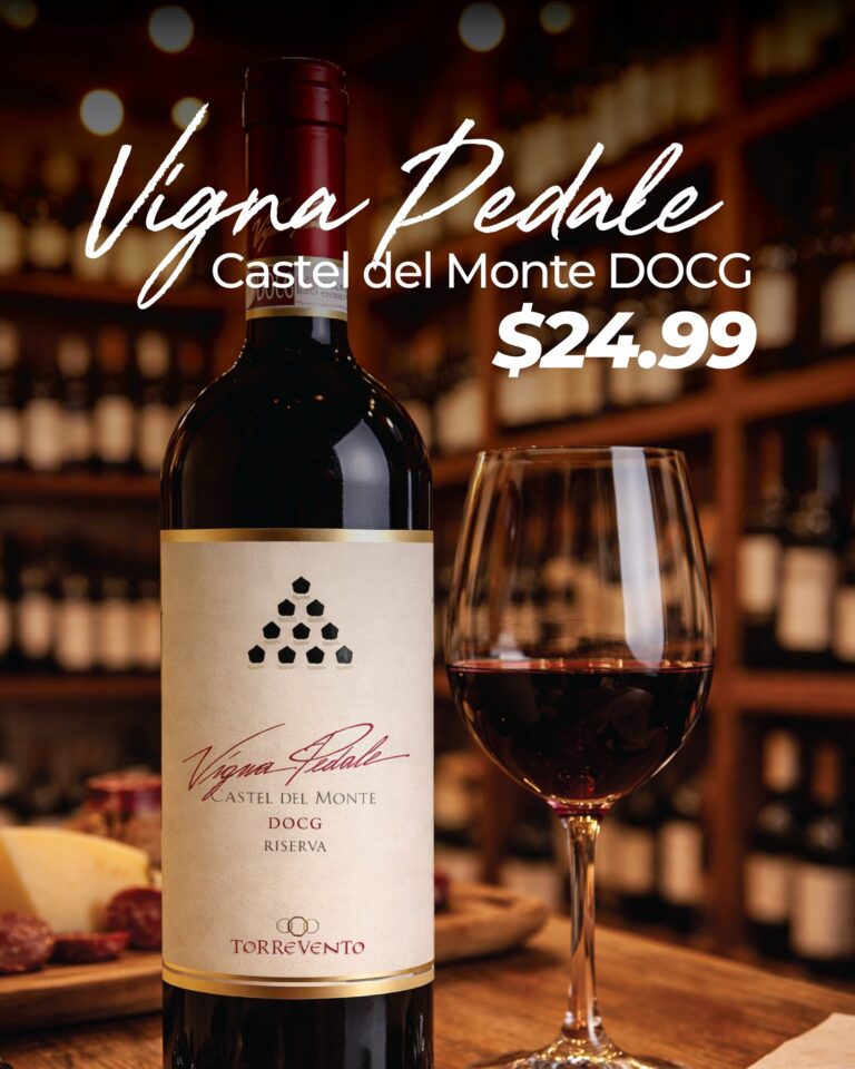 A bottle of Viña Pedale Castel del Monte DOCG Riserva red wine and a filled wine glass are displayed on a wooden surface with a blurred background of wine racks and warm lighting in Roatan, Honduras.