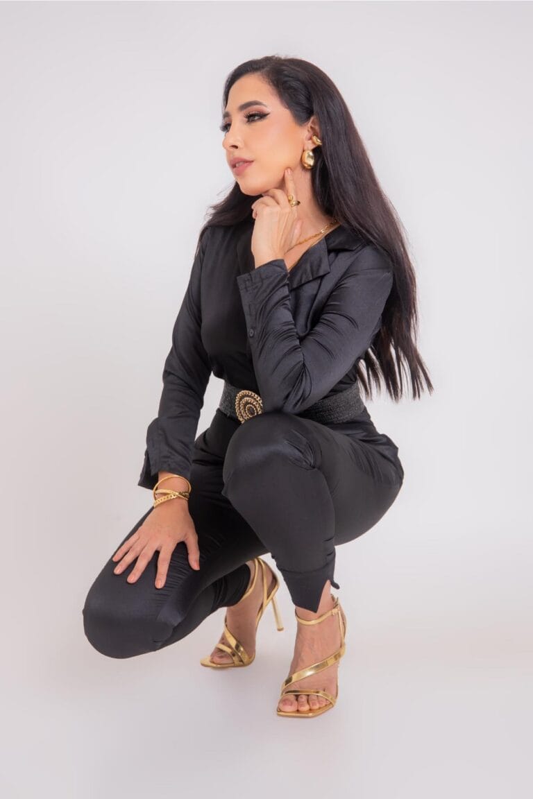 A woman with long dark hair, wearing a black satin blouse and gold high-heeled sandals, poses against a plain white background, highlighting her jewelry, makeup, and fashion style.
