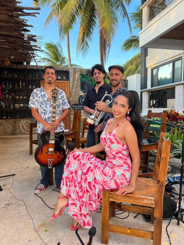 A musical trio performing on an outdoor patio in Roatan, Honduras, features a woman in a pink floral dress sitting on a wooden bench with a microphone, a man with a guitar, and two men holding a trumpet and a microphone, with tropical palm trees and modern buildings in the background.