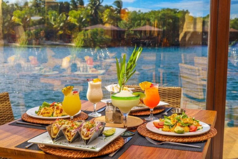 A table set with colorful cocktails, tacos, and salads on a wooden table by a glass window overlooking Roatan's waterfront in Honduras.