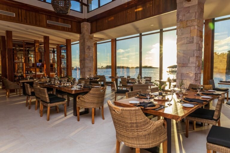 Elegant indoor restaurant in Roatan, Honduras, featuring large glass windows with ocean views, wooden accents, stone pillars, and neatly set tables with black napkins and glassware.