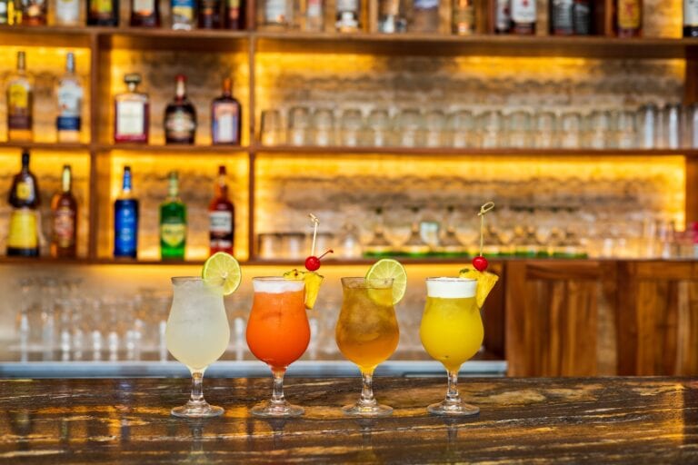 Six colorful cocktails garnished with lime slices and cherries are displayed in front of a wooden bar with liquor bottles and shot glasses in Roatan, Honduras.