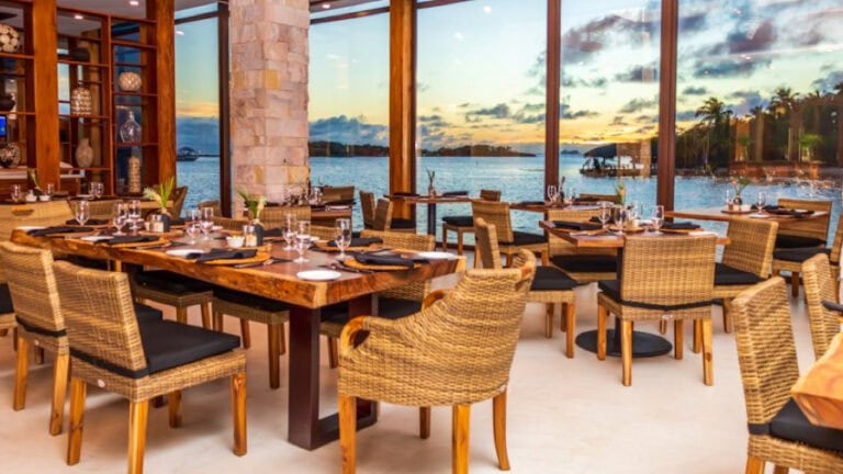 A waterfront restaurant in Roatan, Honduras, features wooden tables set with glassware and black tableware, surrounded by wicker chairs with black cushions, with large glass windows offering a scenic view of the ocean and sunset over the water.