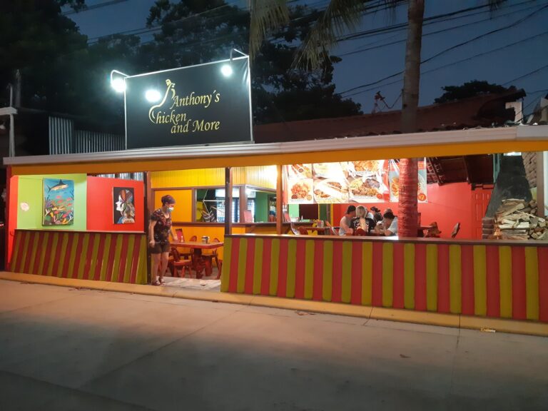 A brightly lit food stand in Roatan, Honduras, features colorful yellow and red striped paneling, with a sign reading "Anthony's Chicken and More" and a group of people dining outdoors at night.