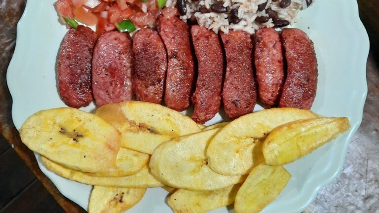 A plate with grilled sausages, fried plantains, fresh tomato and green onion salad, black beans and rice, served on a white dish in Roatan, Honduras.