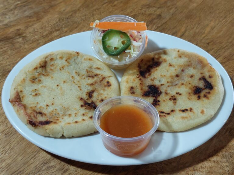 Three arepas served with a side of spicy orange sauce and a small cup of coleslaw topped with jalapeño slice on a white plate on a wooden table in Roatan, Honduras.