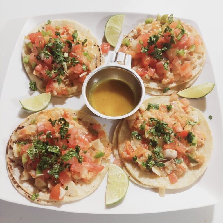 Two fish tacos topped with diced tomatoes, onions, cilantro, and green peppers served with lime wedges and a small container of salsa on a white rectangular plate in Roatan, Honduras.