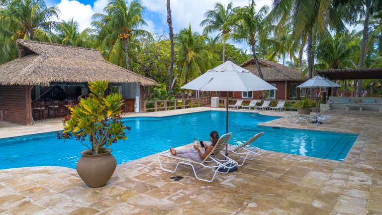 A woman relaxes on a lounge chair by a swimming pool surrounded by palm trees and thatched-roof cabanas on Roatan, Honduras.