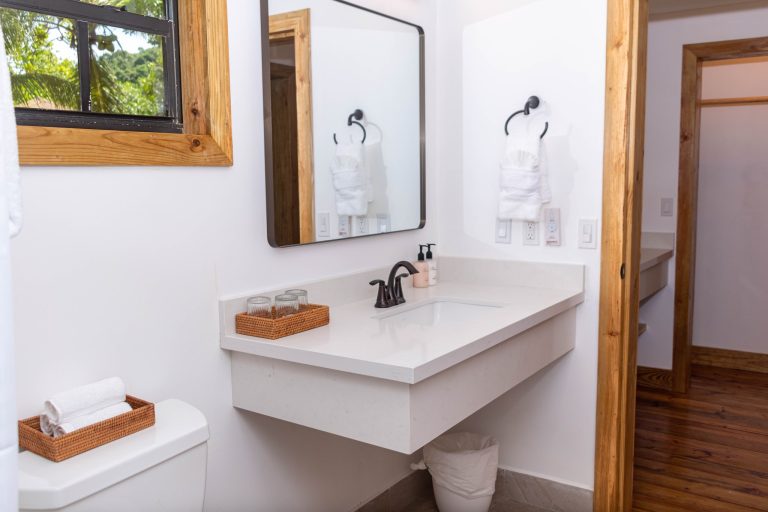 A clean, modern bathroom in Roatan, Honduras, featuring a white vanity with a black faucet, a wall-mounted mirror, a white towel with a towel ring, and a small woven basket holding glassware, with natural wooden door and window frames and a view of green foliage outside.