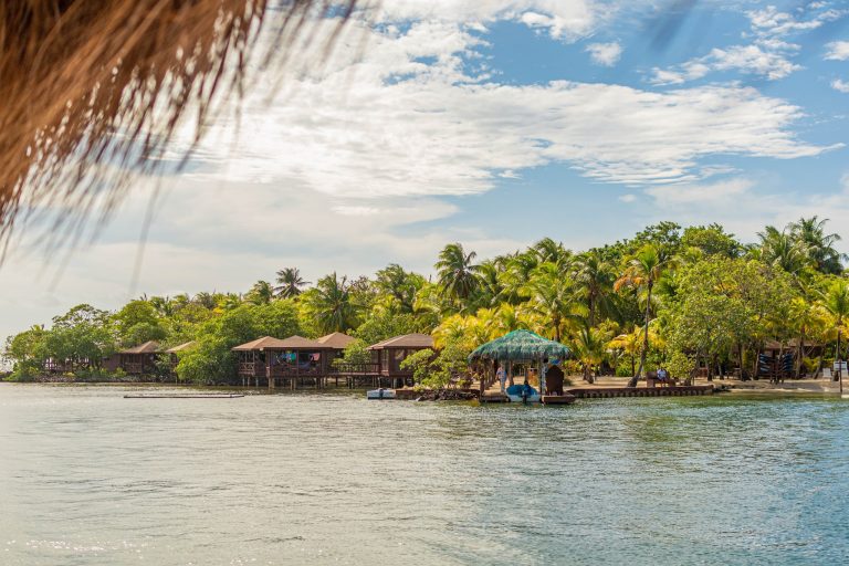Overwater bungalows with thatched roofs are set among lush green palm trees along the shoreline on Roatan, Honduras, under a partly cloudy sky.