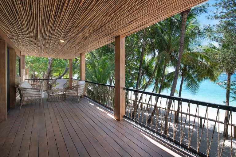 A wooden balcony overlooking palm trees and a turquoise beach in Roatan, Honduras, with wicker chairs and a view of the ocean under a clear blue sky.