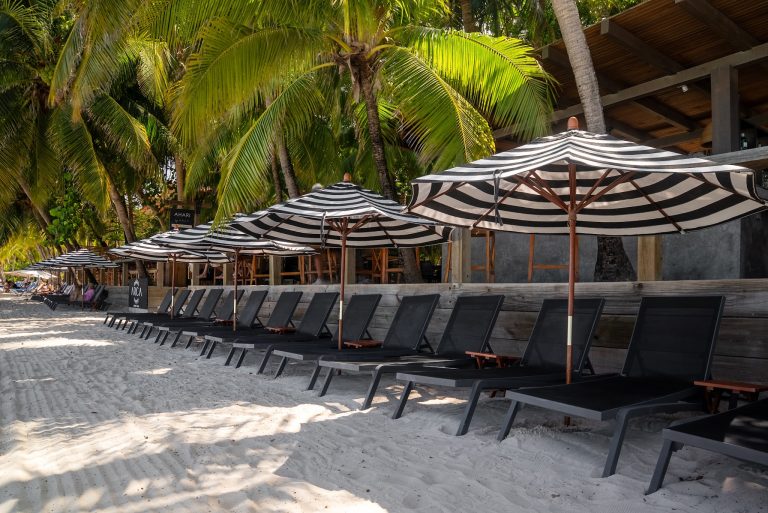 Beach chairs and black-and-white striped umbrellas line the sandy shore beneath lush green palm trees at a seaside location in Roatan, Honduras.