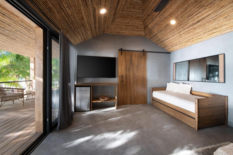 A modern bedroom with a wooden bed and sliding glass door opening to a outdoor balcony surrounded by lush greenery in Roatan, Honduras.