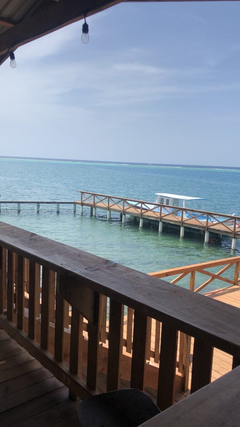 A wooden deck with railing overlooks clear turquoise waters and a small pier extending into Roatan, Honduras, under a blue sky.