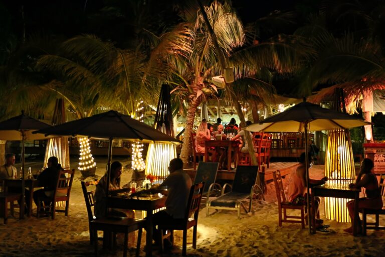 A lively beachside restaurant in Roatan, Honduras, is illuminated with string lights, featuring wooden tables, umbrellas, and palm trees, with people enjoying their evening dining on the sandy shore.