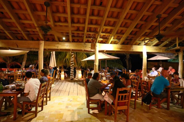 A lively dining area at a restaurant or bar in Roatan, Honduras, featuring wooden furniture, string lights, palm trees, and patrons enjoying their meals outdoors at night.