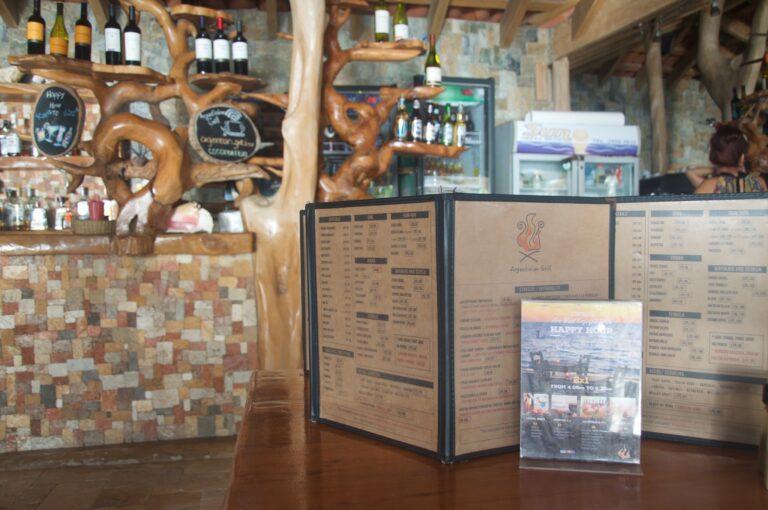 A wooden interior dining area in Roatan, Honduras features a rustic stone wall, a menu display, and decorative wooden branches, with a bar in the background stocked with beer bottles and a woman sitting at the counter.